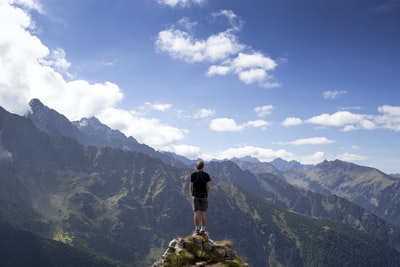 在白色和蓝色多云的天空下，穿着黑色衬衫和灰色短裤的男子在山旁的山上